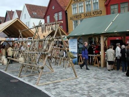 Tørrfiskhjeller av rundstokk reist på brosteinen foran de gule trebygningene på Bryggen, med festivaltelt og publikum