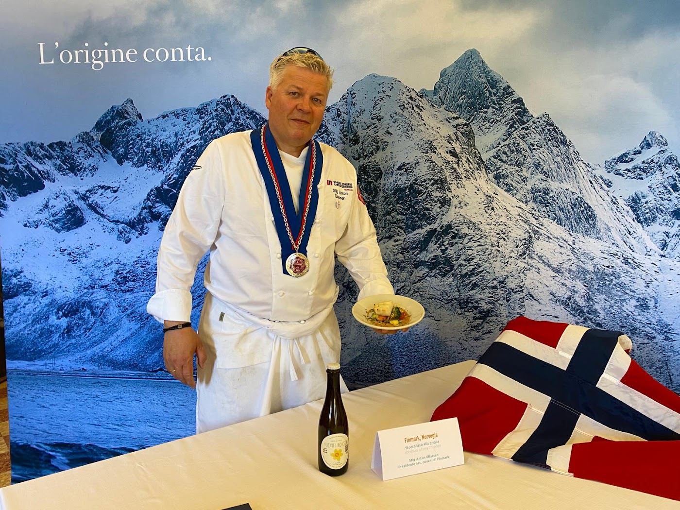 Kokk i hvit kokkejakke med ordenskjede holder fram en tallerken med tørrfiskrett foran en plakat med lofotfjell og norsk flagg