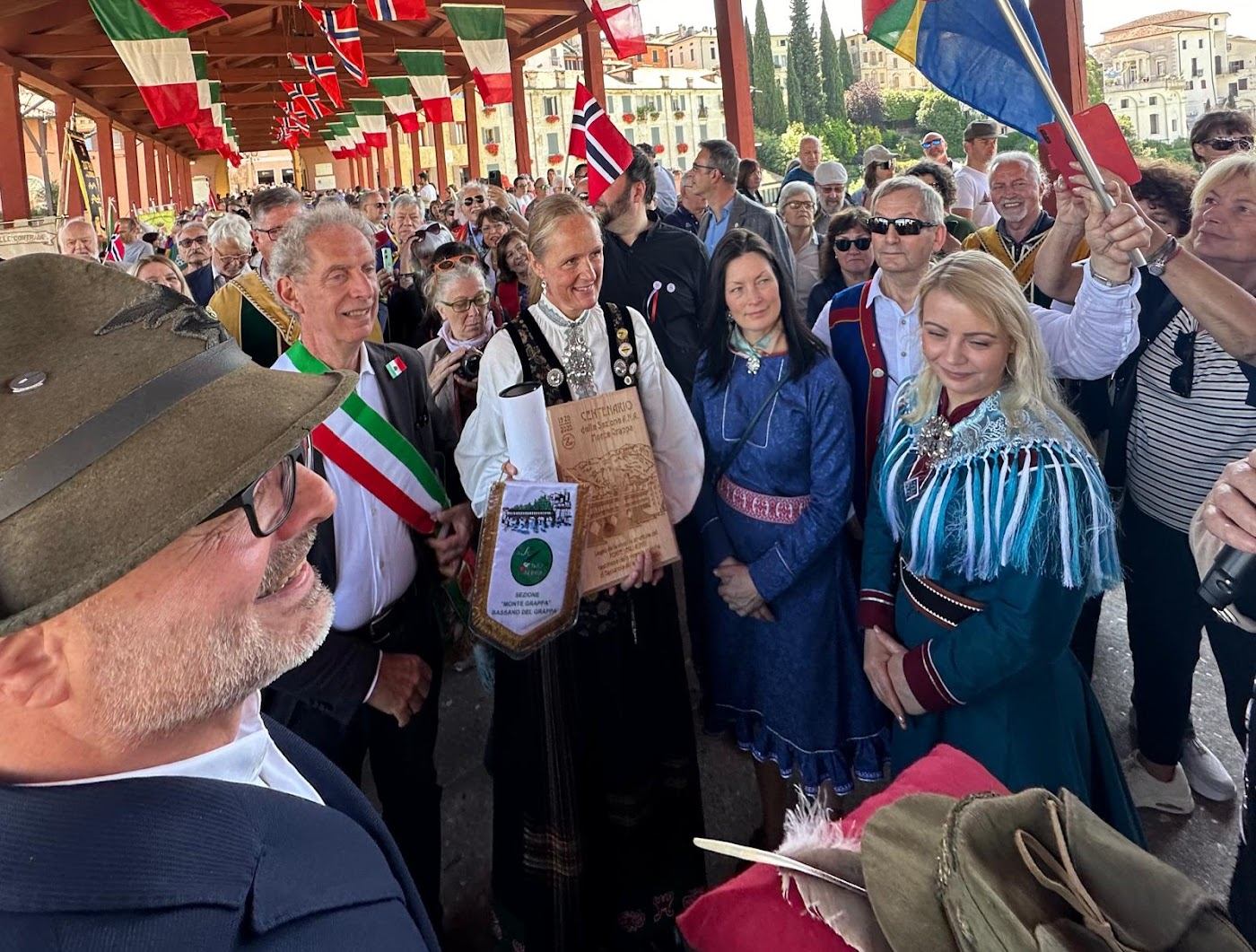Folkemengde under italienske, norske og samiske flagg på den overbygde broen; en kvinne i bunad holder en utskåret treplakett, og en alpinihatt med lang fjær ligger på en rød pute i forgrunnen