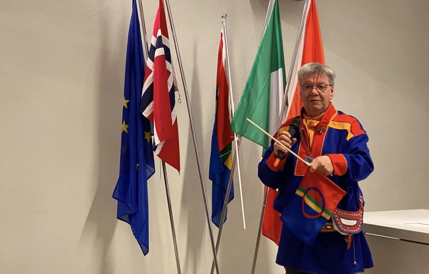 Mann i blå og rød samisk gákti holder mikrofon og et lite italiensk flagg foran en rekke flaggstenger med EU-, norsk, samisk og italiensk flagg