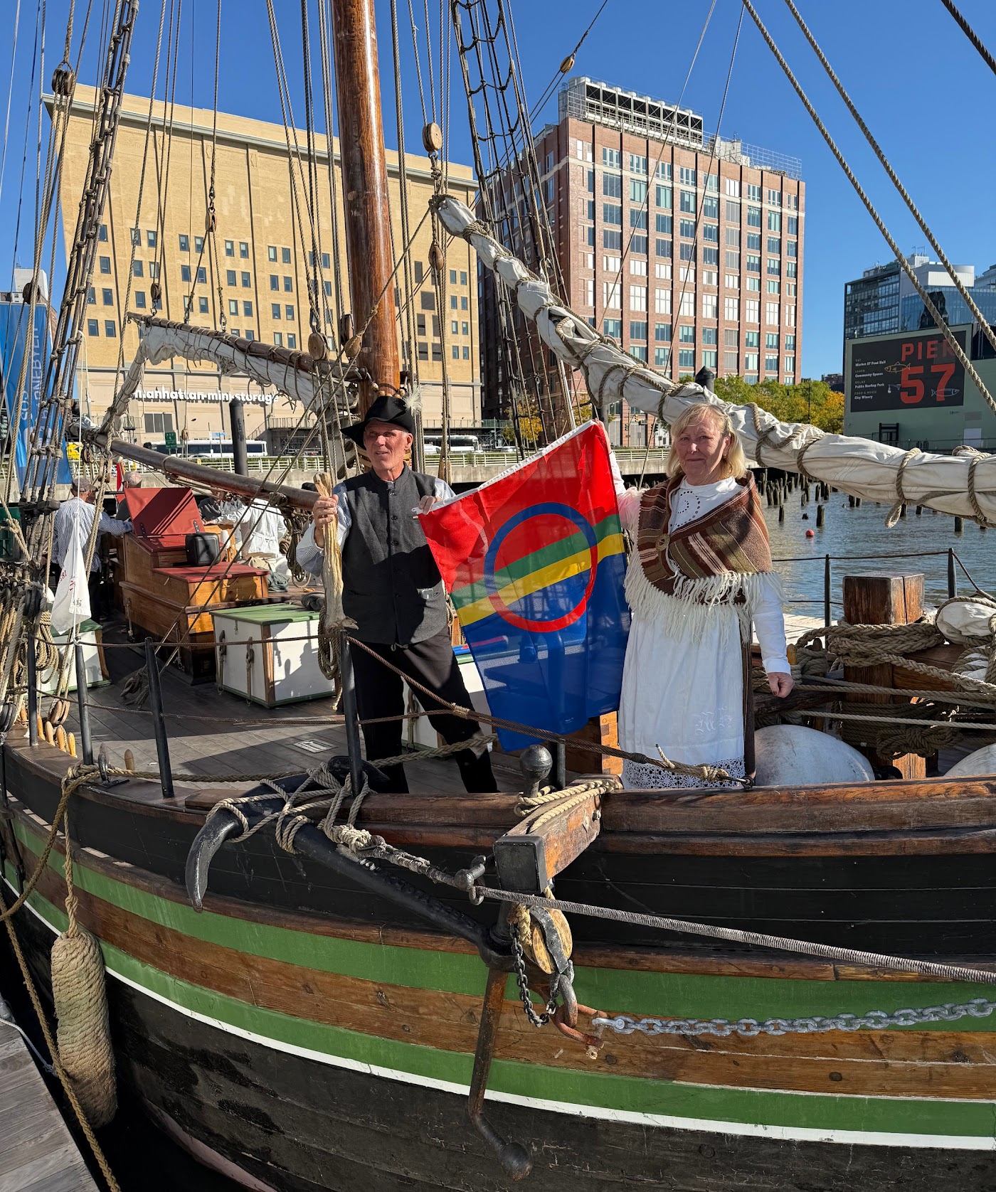 The sailing sloop Restauration moored at Pier 57 in New York; a man and a woman in historic costume hold up the Sámi flag.