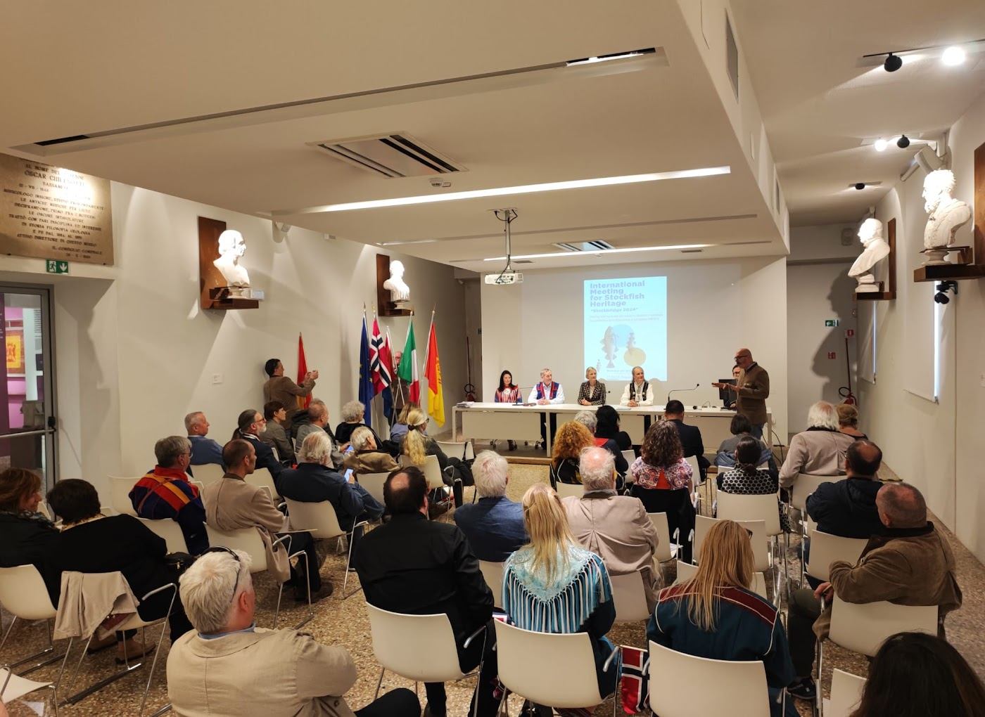 Konferansesal i Bassano med lerret merket «International Meeting for Stockfish Heritage – Stockbridge 2024». Panel i samiske og norske drakter, flagg fra fire land langs veggen.