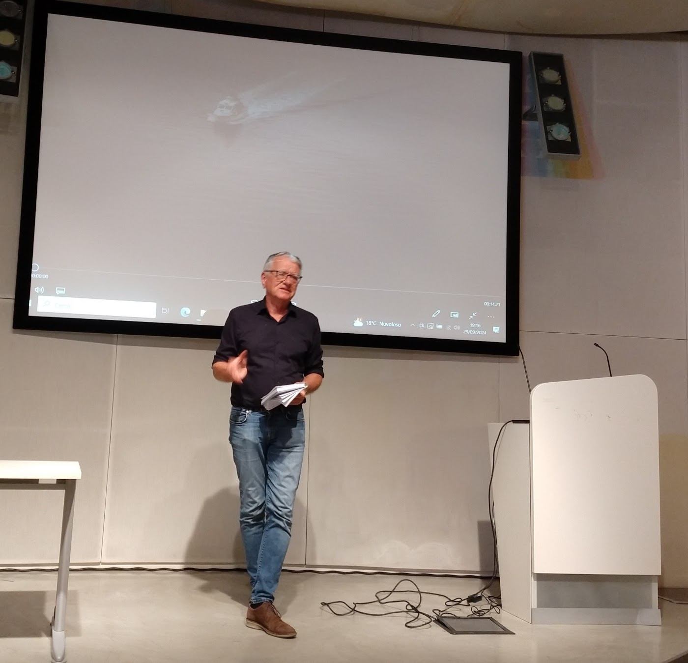 A man stands in front of a large projection screen presenting a film to an audience.