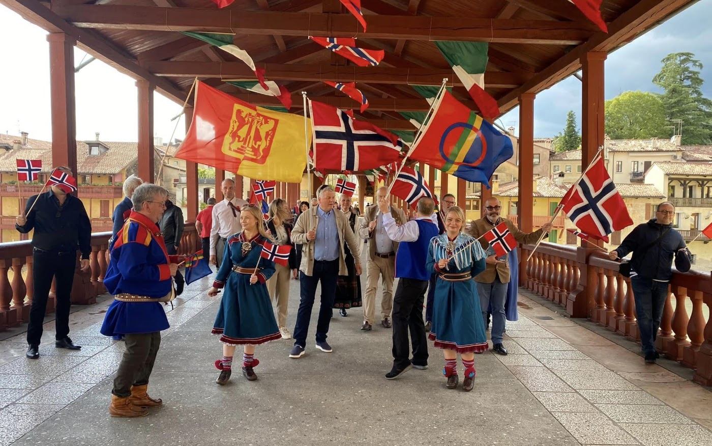 Deltakere i samisk gákti og norsk bunad bærer norske og samiske flagg i prosesjon gjennom den overbygde trebroen i Bassano del Grappa