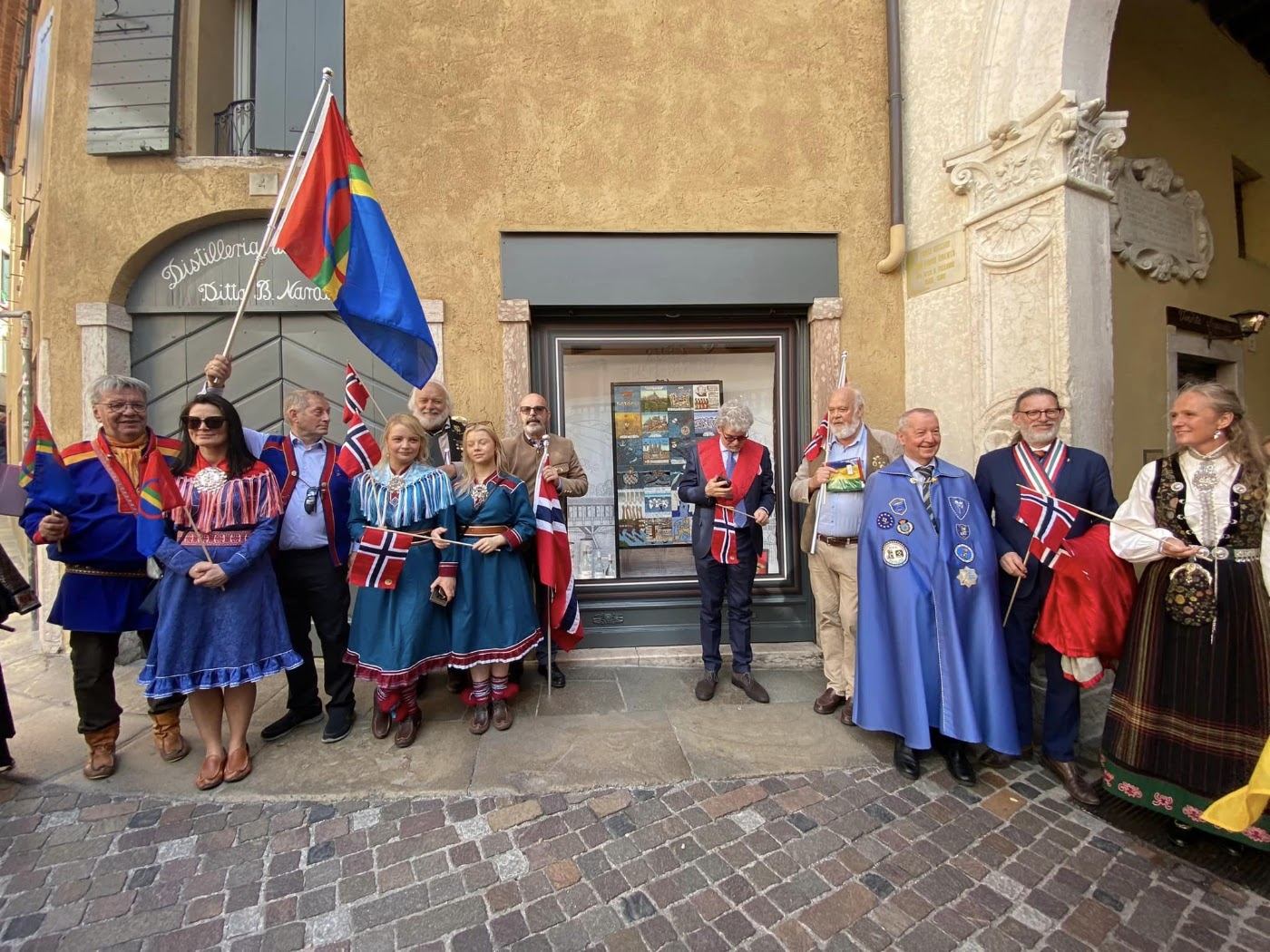 Mennesker i samiske kofter, bunad og italienske brorskapskapper står med norske og samiske flagg foran et butikkvindu med utstilte keramiske fliser i Bassano del Grappa