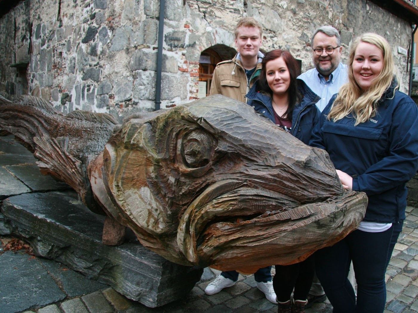 Fire personer står bak en stor treskulptur av et torskehode som ligger på steinheller foran en gammel steinmur på Bryggen i Bergen
